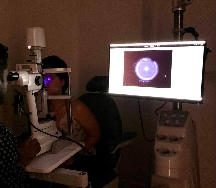 Port Chester Eye Care doctor is examining a patient’s eyes using a slit lamp. The patient is sitting in front of the machine, and the doctor is looking through the eyepiece. The patient’s chin is resting on the chin rest, and the doctor is using a joystick to adjust the position of the light. The room is dimly lit, and there are other medical instruments visible in the background.